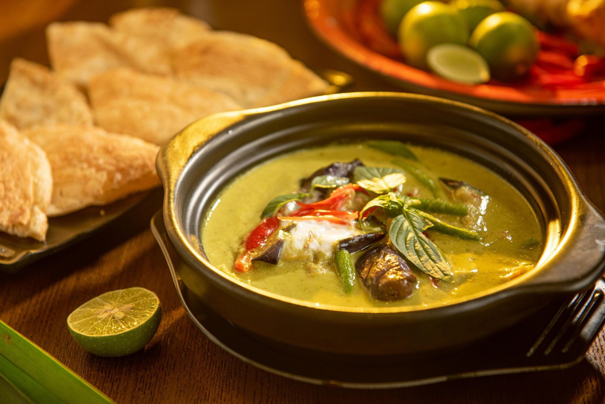 A hearty bowl of creamy green curry features chunks of eggplant, red chili slices, and a garnish of fresh basil leaves. The dish is elegantly presented on a wooden table alongside a plate of golden flatbread and a fresh lime wedge.