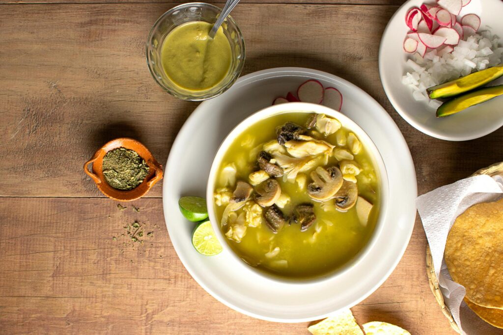 A bowl of green pozole containing mushrooms and hominy sits on a rustic wooden table, served with fresh lime wedges and a small clay dish of dried herbs. The meal is accompanied by traditional sides, including a basket of tostadas, a bowl of green salsa, and a plate of sliced radishes, onions, and avocado.