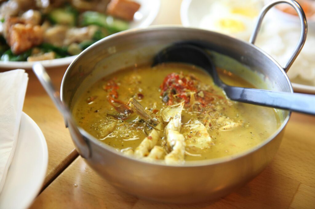 A stainless steel bowl contains a yellow seafood curry garnished with red chilies, with a black ladle resting inside for serving. The dish is placed on a wooden table amidst other plates, including stir-fried vegetables and boiled eggs visible in the background.