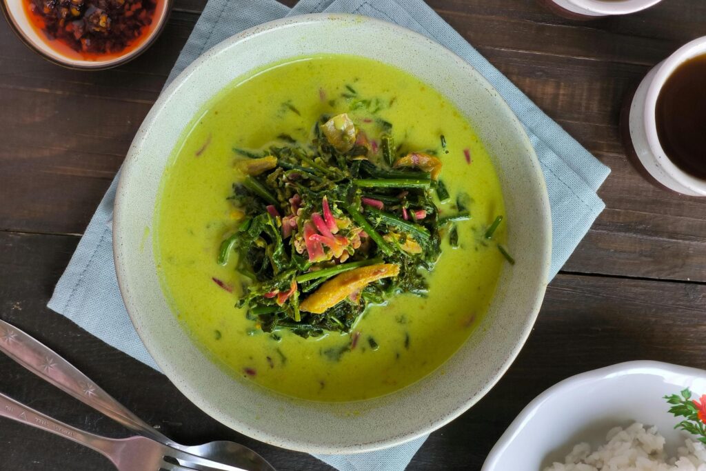 A ceramic bowl presents a creamy, light green curry centered with a mound of dark green vegetables and pink aromatic garnishes, resting on a light blue napkin. The setting includes silver cutlery to the left, a plate of white rice to the right, and a small side dish of dark red chili paste.