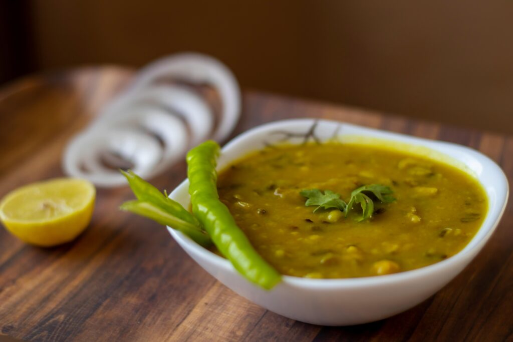 A white bowl holds a thick yellow lentil curry (Dal), garnished with fresh cilantro and a whole green chili pepper resting on the rim. The dish is presented on a wooden board accompanied by sliced onion rings and a lemon half, adding zest and crunch to the meal.