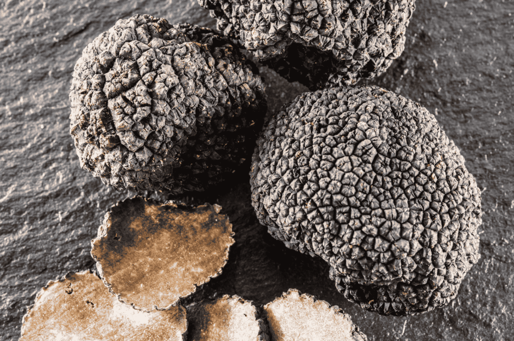 Black truffles arranged on a dark, textured surface, with a few thin slices revealing their marbled interior. The composition emphasizes the fungi’s rugged exterior and aromatic complexity, echoing Piedmont’s culinary reverence and seasonal abundance.