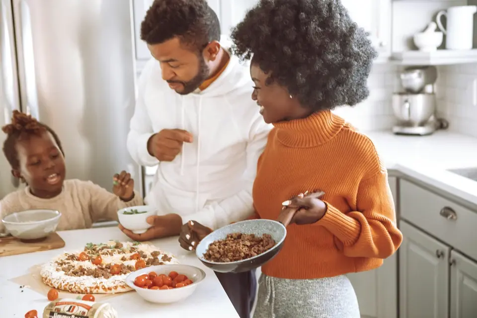 A happy family of three—a man, a woman, and a young girl—standing in a bright kitchen assembling a homemade pizza with fresh toppings.