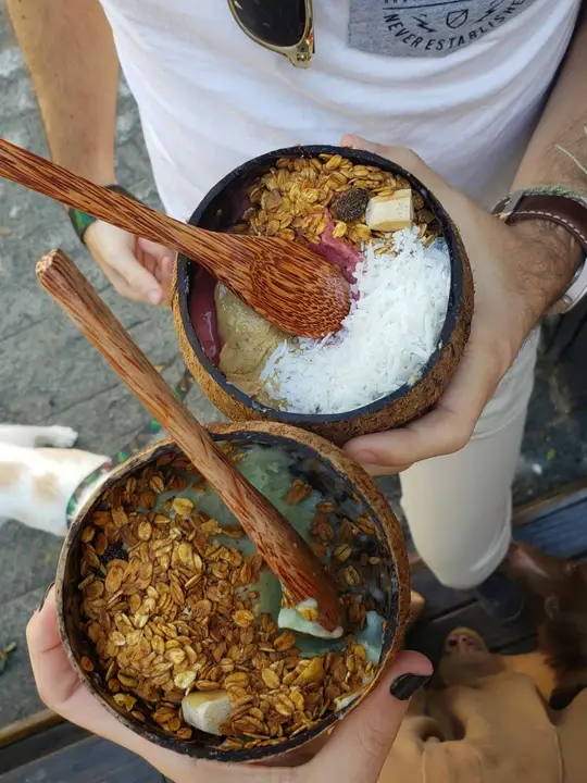 A top-down view of two people holding coconut shell bowls filled with vibrant smoothie bases, topped with granola, shredded coconut, and sliced fruit.