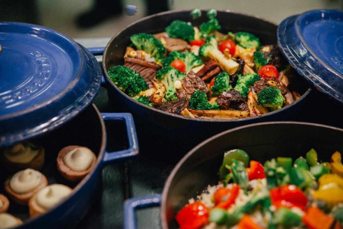 Several blue enameled cast iron pots filled with colorful prepared meals, including a stir-fry with broccoli, beef, and cherry tomatoes.