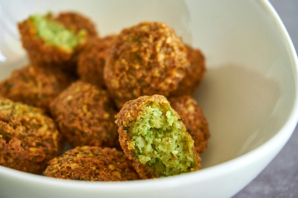 A white bowl is filled with golden-brown, crispy falafel balls, one of which is broken open to reveal a vibrant green, textured center. The shallow depth of field keeps the foreground falafel in sharp focus while the others softly blur into the background.