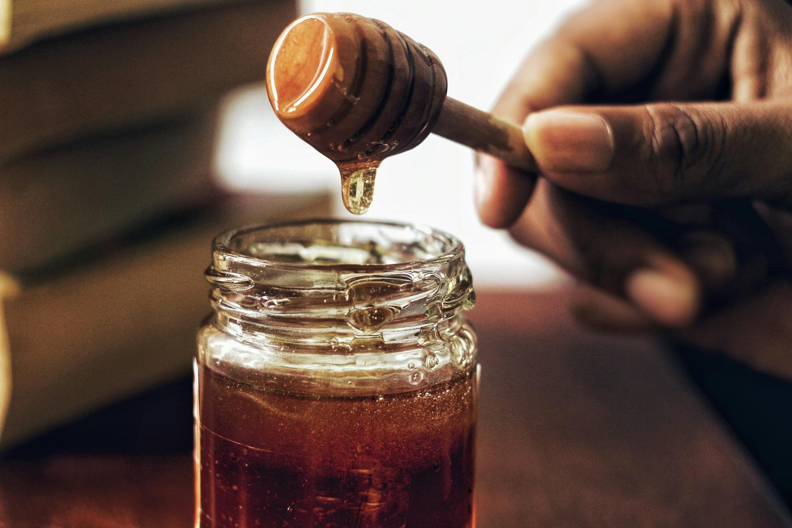 A hand holds a wooden honey dipper over an open glass jar, catching a single golden drop as it falls. The warm, amber-toned scene is set against a soft-focus background of stacked books.