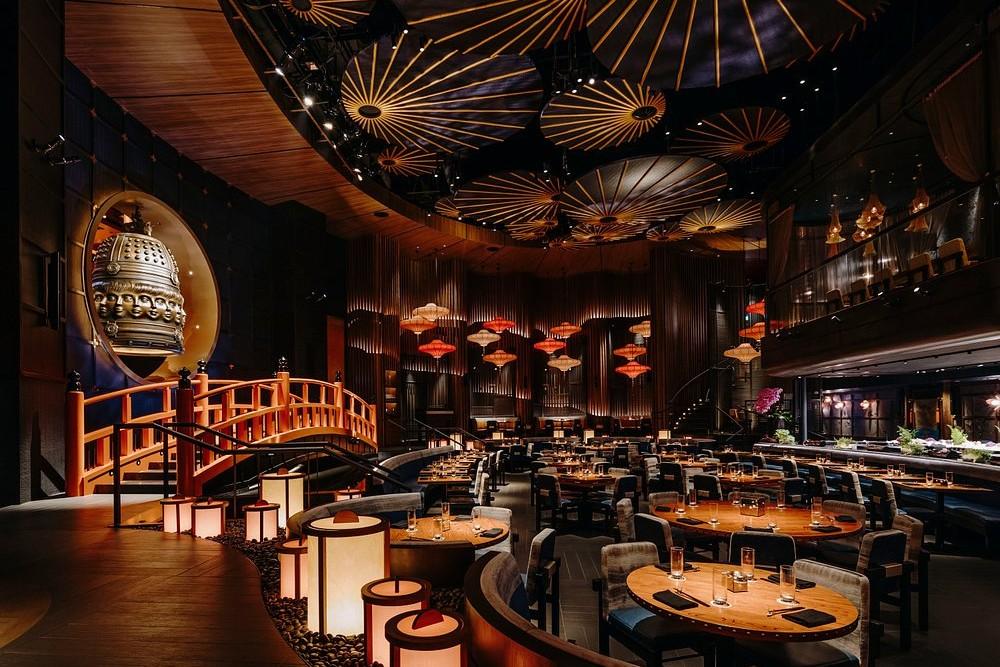 This image captures the opulent, high-ceilinged main dining room of KOMA Singapore, featuring a dramatic Japanese-inspired interior with a traditional orange footbridge and a massive bronze bell. The space is atmospheric and warm, illuminated by glowing lanterns, circular ceiling fans that resemble umbrellas, and rows of sleek, circular wooden tables.