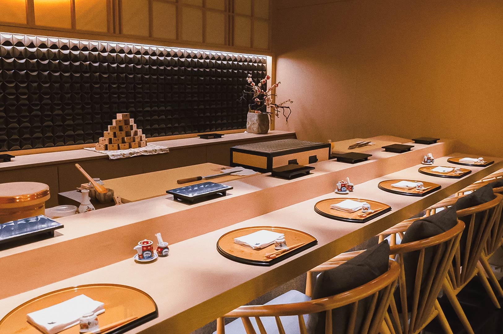 Japanese omakase sushi bar interior featuring lacquered trays, chopsticks, sake cup display, and flower arrangement.