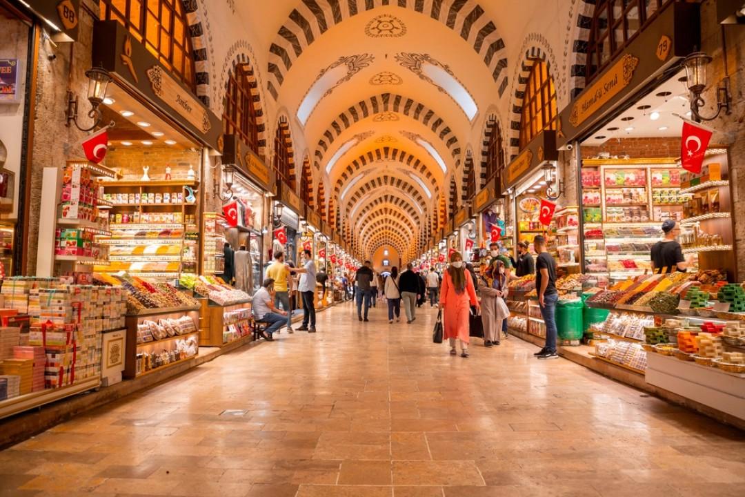 This image captures a vibrant, bustling walkway inside the Spice Bazaar in Istanbul, featuring high vaulted ceilings with distinctive striped arches. Shoppers stroll past numerous stalls filled with colorful mounds of spices, sweets, and traditional goods, all under the glow of warm overhead lighting.