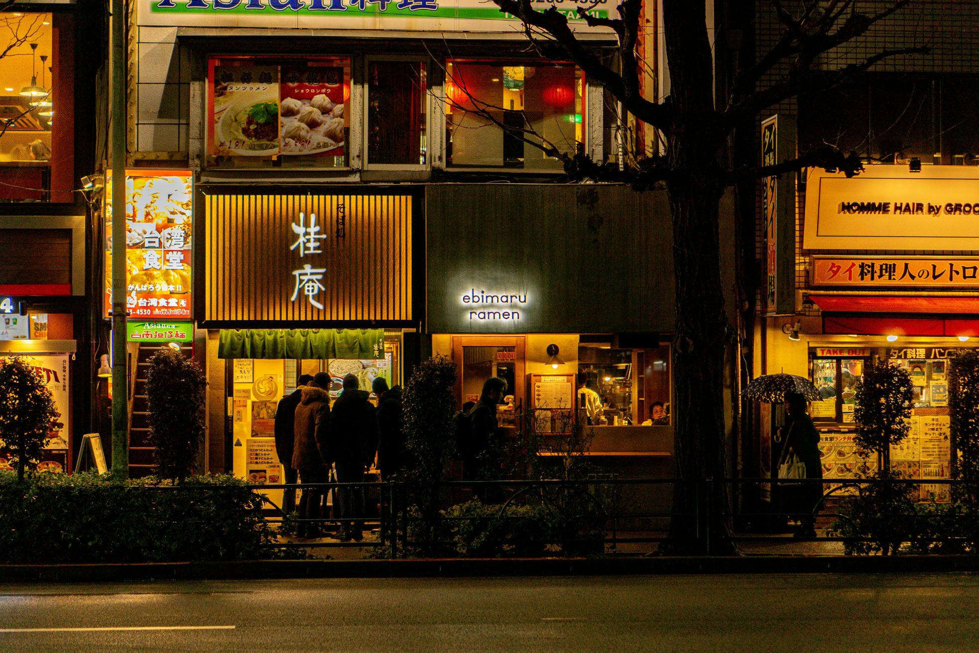 This nighttime street scene in Japan features a row of small restaurants, including Ebimaru Ramen, glowing with warm yellow and orange light. A group of people stands on the sidewalk near the shop entrances, framed by dark trees and colorful signage.