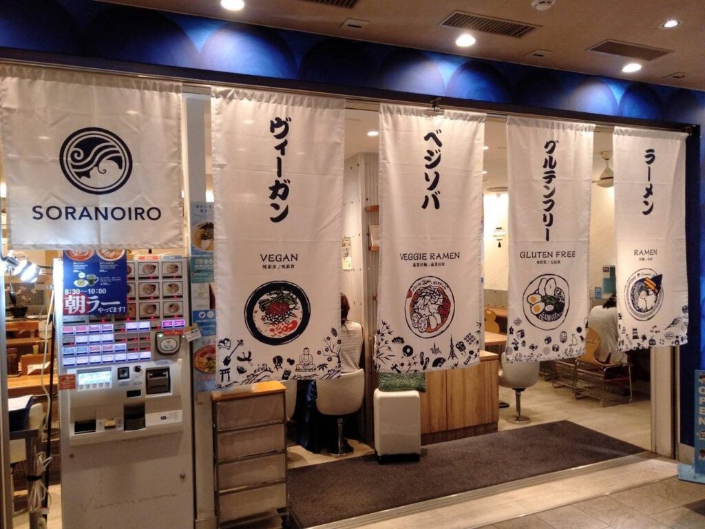 The entrance of this Soranoiro restaurant is adorned with long white banners advertising vegan, veggie, and gluten-free ramen options. Beside the doorway, a ticket vending machine with numerous illuminated buttons is available for customers to place their orders.