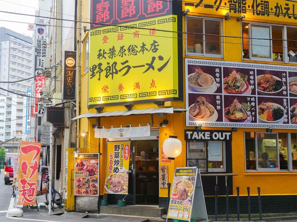 The exterior of this Yarou Ramen shop is painted a vibrant yellow and features large signs with photos of various ramen bowls. A small take-out window is situated next to the entrance, which is decorated with posters and a standing menu board on the sidewalk.