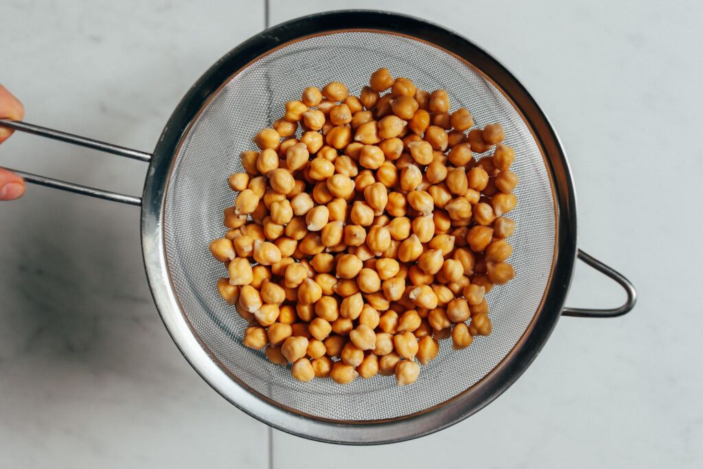 A metal mesh strainer filled with cooked, golden-brown chickpeas is held over a light-colored countertop. The chickpeas appear moist and tender, ready to be used in a recipe or served.