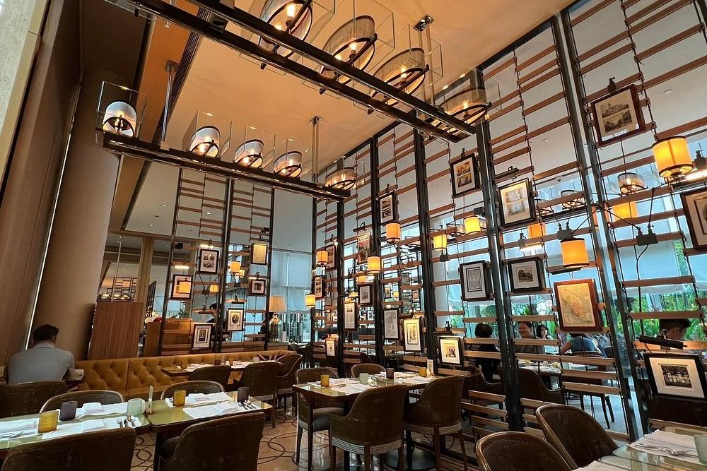 This restaurant interior features a sophisticated design defined by soaring ceilings and high, lattice-style partitions adorned with framed artwork and glowing amber lanterns. The dining area is furnished with tufted banquettes and wicker-backed chairs arranged around set tables, all under the warm light of large, glass-encased rectangular chandeliers.