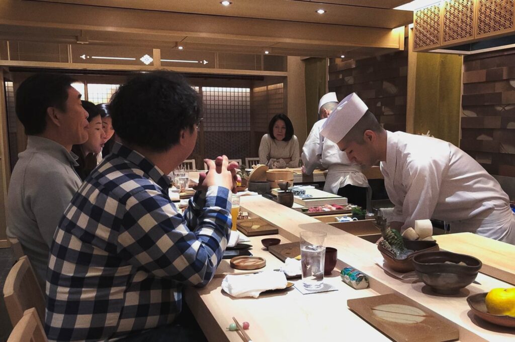 Traditional Japanese omakase sushi restaurant interior with chefs in white uniforms preparing fresh sushi at wooden counter for seated patrons.