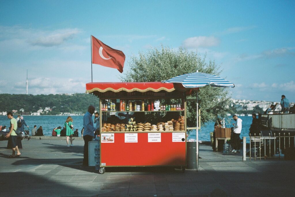 A red street food cart topped with a Turkish flag stands prominently on a sunny waterfront, displaying stacks of traditional simit and bottled drinks. People stroll along the sunlit promenade in the background, with the blue waters of the Bosphorus and distant hills framing the scene.