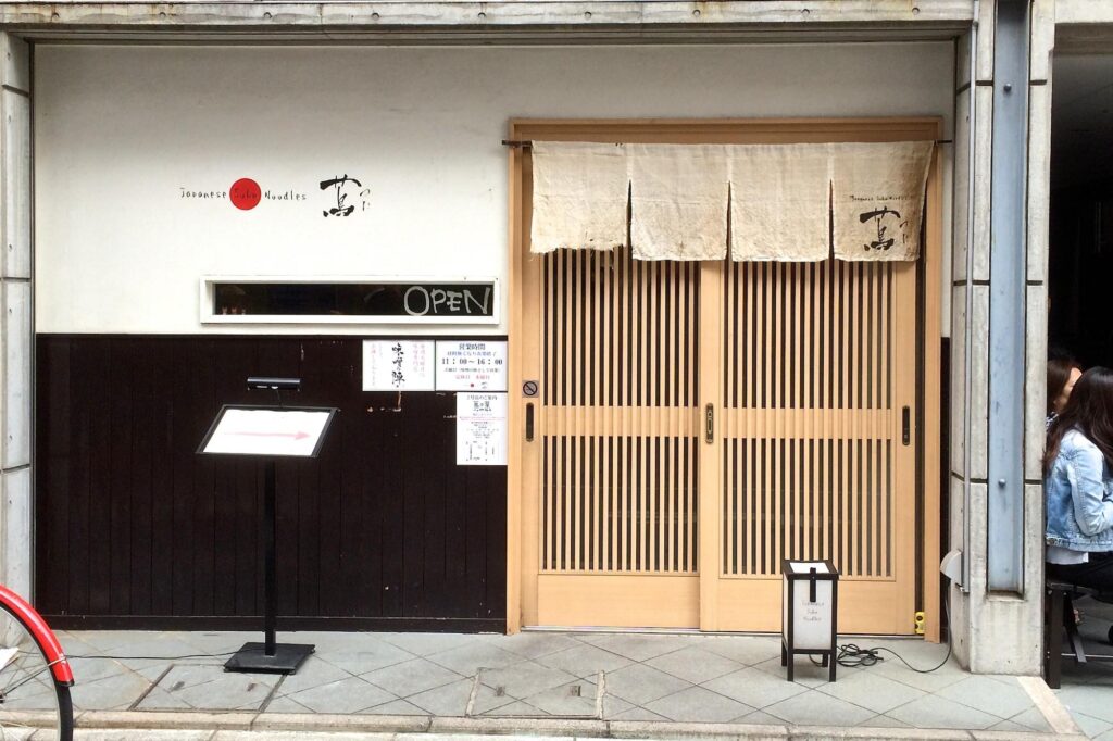 This image shows the minimalist storefront of a Japanese Soba Noodles restaurant featuring a traditional wooden slatted door and a cream-colored noren curtain. The exterior is composed of white and dark wood paneling, accented by a small "OPEN" sign and a standing menu pedestal on the sidewalk.