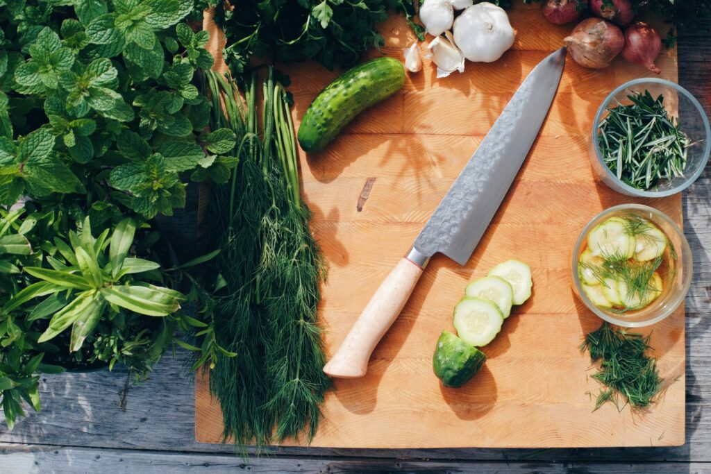 A chef's knife rests on a wooden cutting board surrounded by fresh ingredients like cucumbers, dill, garlic, and mint. Several cucumber slices and chopped herbs are spread across the board alongside small bowls of prepared garnishes.