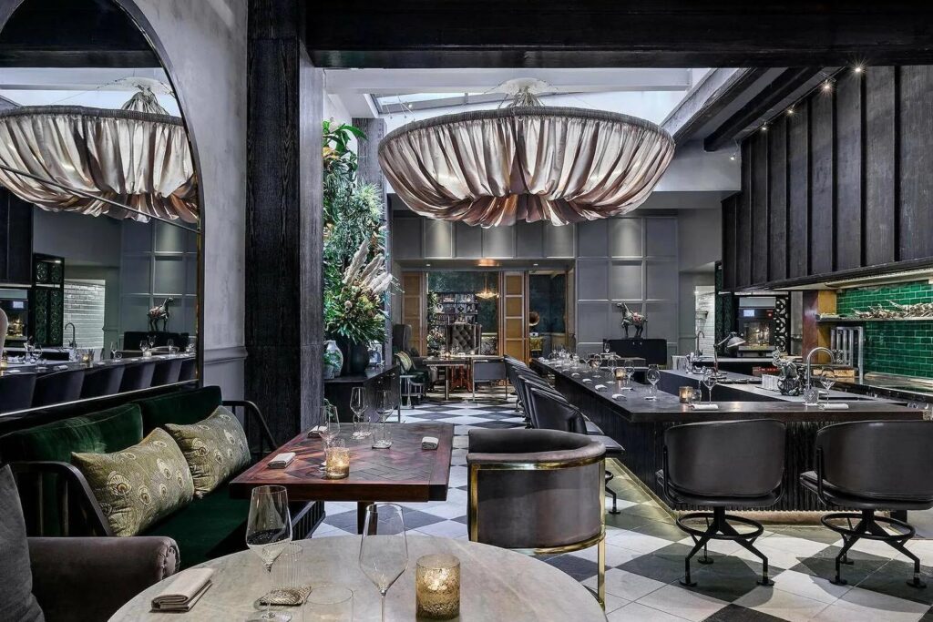 The dining room showcases an eclectic, upscale design highlighted by a large, draped fabric chandelier hanging above a checkerboard floor. Dark wood accents and velvet seating create a moody atmosphere, while an open kitchen area with green tiles and a tall floral arrangement add pops of color to the space.