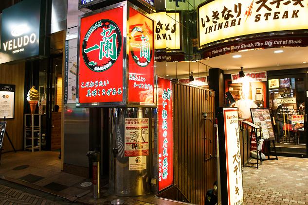 Brightly lit signs for Ichiran Ramen and Ikinari Steak dominate this vibrant nighttime street corner in Japan. The bustling scene features a mix of glowing red and yellow advertisements alongside a coffee shop and restaurant entrances.