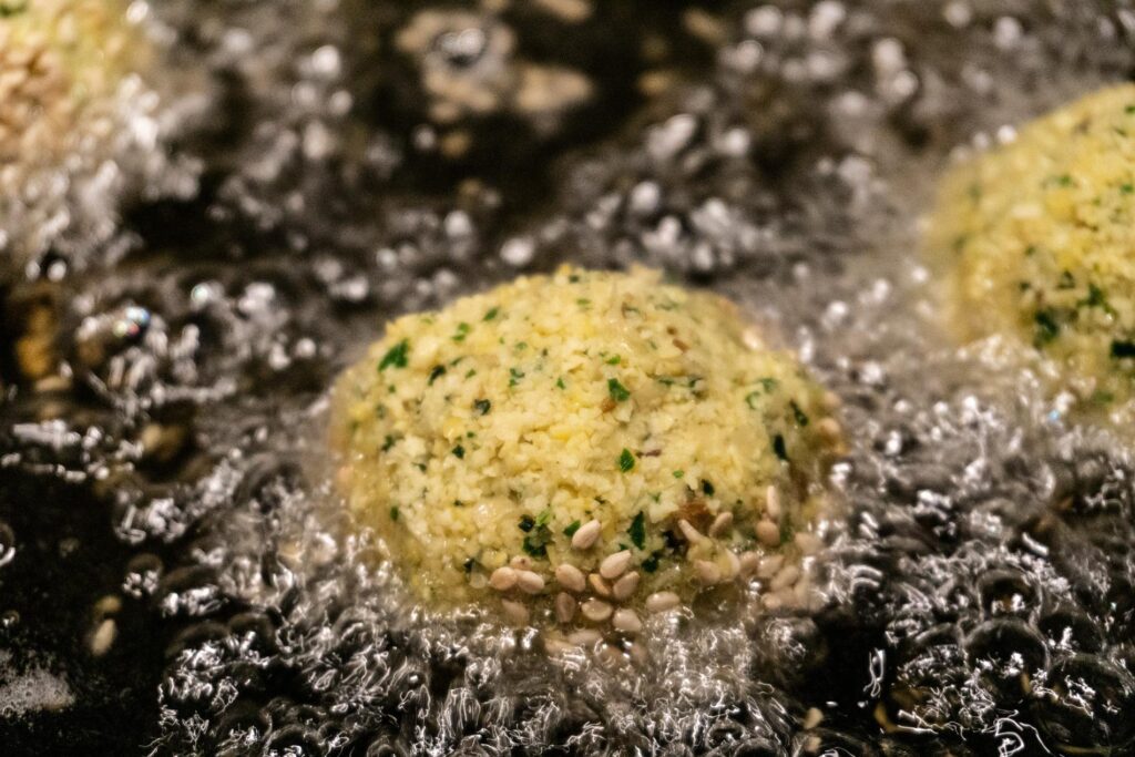 Falafel balls are shown submerged in hot, bubbling oil during the deep-frying process. The centered falafel features a light yellow, herb-flecked interior and is coated with several visible sesame seeds.