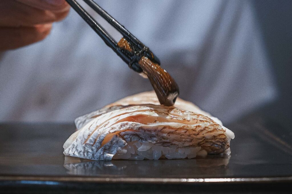Japanese omakase sushi close-up showing chef brushing soy-based sauce onto white fish nigiri with chopsticks.