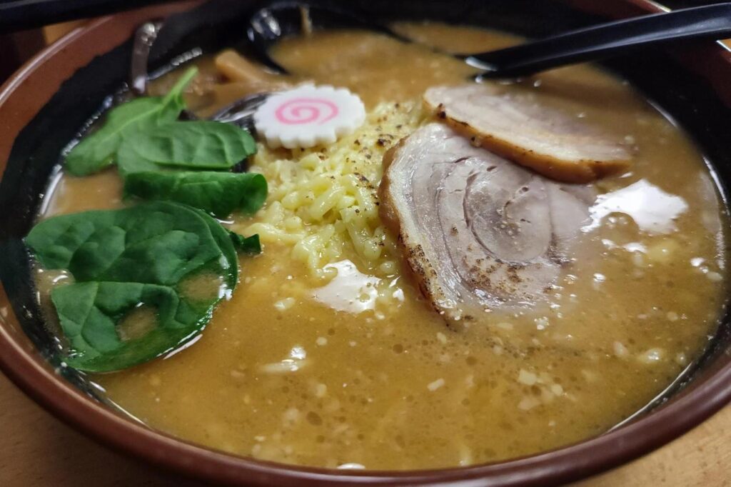 A steaming bowl of miso cheese ramen is topped with thick slices of chashu pork, fresh green leaves, and a pink-swirled narutomaki fish cake. The rich, savory broth surrounds the noodles and toppings, with black spoons resting along the edge of the dark bowl.