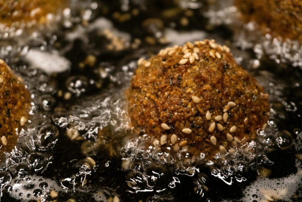 Several falafel balls are being deep-fried in bubbling oil, with one prominently centered and covered in sesame seeds. While the surrounding oil vigorously fizzes, the falafel's exterior has developed a rich, dark golden-brown crust.