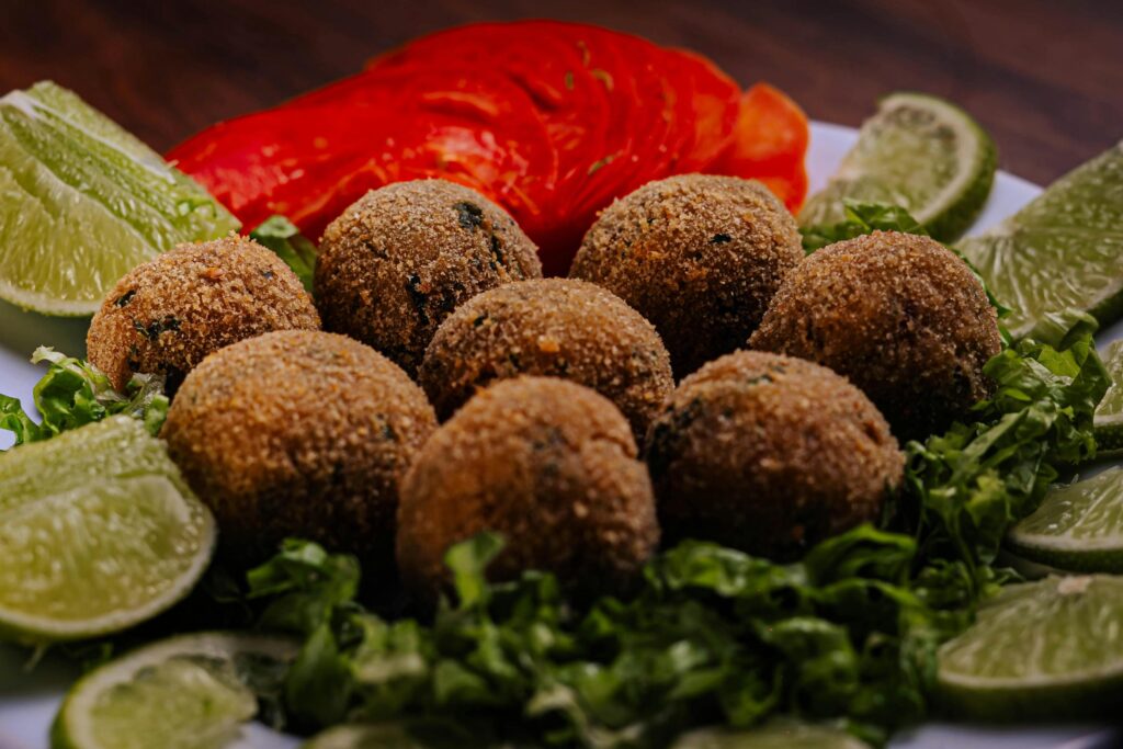 A plate features several golden-brown, breaded falafel or croquette balls nestled on a bed of shredded green lettuce. The dish is garnished with bright lime wedges and thinly sliced red tomatoes, adding a vibrant pop of color to the presentation.