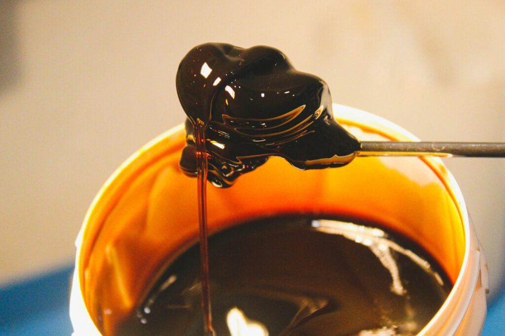 A metal tool lifts a thick, glossy glob of dark amber honey from a white container. The viscous liquid stretches into a smooth, translucent thread as it drips back into the pool below.