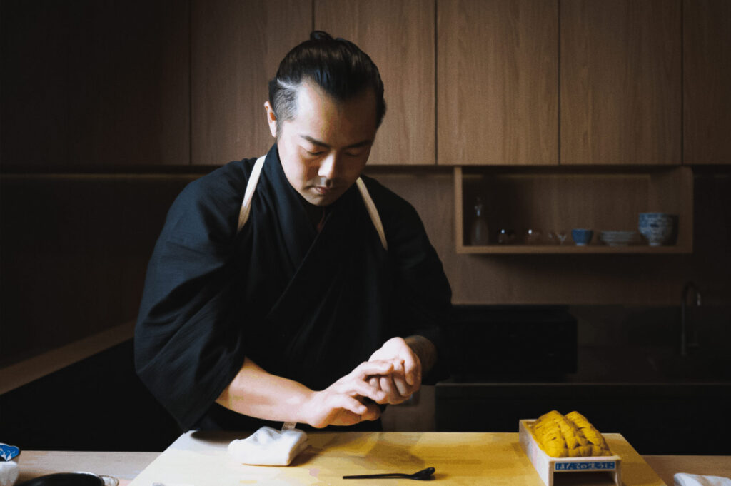 Sushi Masa omakase chef preparing uni nigiri with fresh sea urchin in wooden box at modern sushi counter.