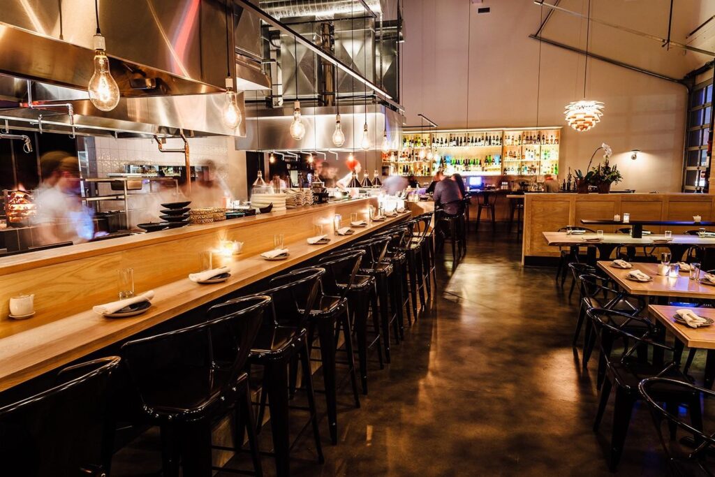 This modern, high-ceilinged restaurant features a long wooden counter with black barstools and an open kitchen where chefs are visible behind hanging industrial lights. The spacious interior is warmly lit, showcasing a well-stocked bar in the background and several dining tables neatly arranged for guests.