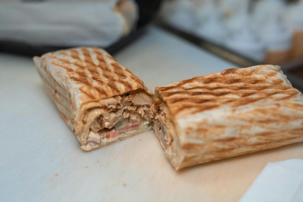 A grilled wrap is sliced in half to reveal a filling of seasoned meat, fresh tomatoes, and creamy sauce. The golden-brown exterior features distinct grill marks, indicating it was pressed for a crispy texture.