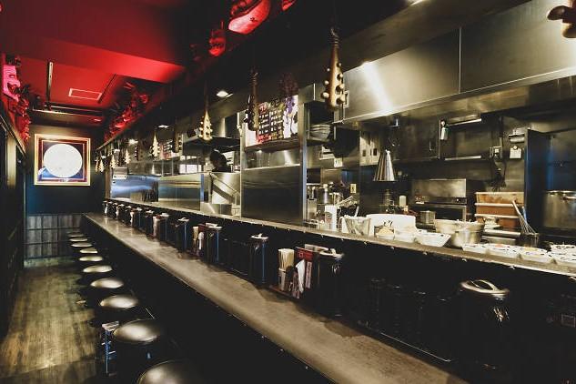 This narrow, dimly lit restaurant features a long, dark dining counter lined with round black stools under a striking red ceiling. The industrial-style open kitchen is visible behind the counter, accented by unique club-shaped ornaments and red masks hanging from the walls.