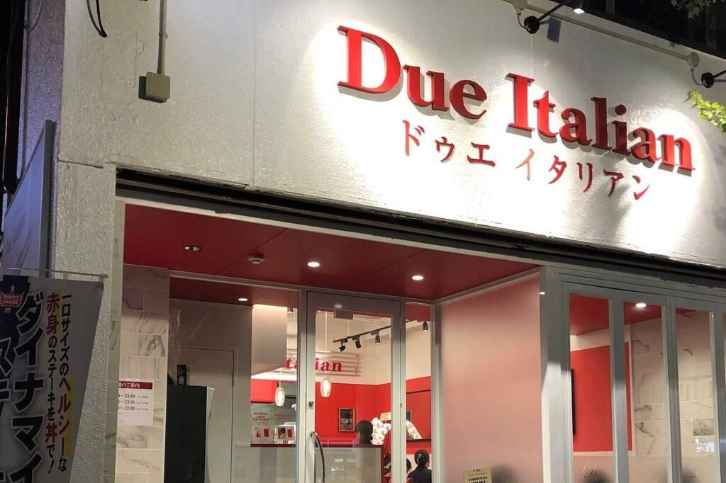 The storefront of "Due Italian" features a bright white facade with large red lettering and a clean, modern entrance. Through the glass doors, the interior is visible with a striking red ceiling and minimalist decor that matches the restaurant's branding.