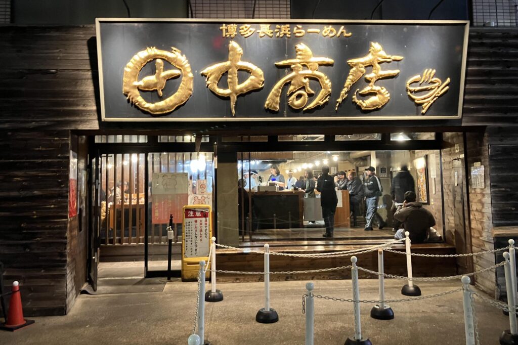 This restaurant storefront features a large, dark signboard with prominent gold-colored Japanese characters and a logo of a steaming bowl of noodles. The building has a rustic dark wood exterior, and through the large windows, customers are visible eating and standing inside the brightly lit dining area.