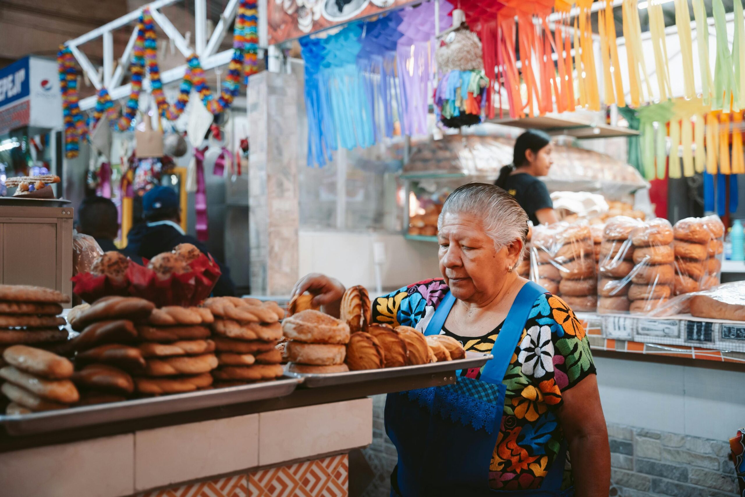 An elderly woman wearing a vibrant, floral-patterned shirt and a blue apron carefully arranges a variety of traditional Mexican pan dulce on a metal tray at an outdoor market. In the background, colorful paper decorations and garlands hang above the stalls, where another worker and stacks of bagged bread are visible.