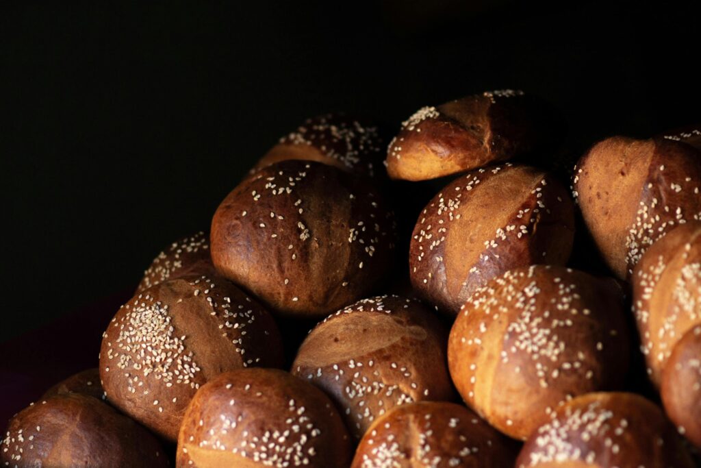 A pile of golden-brown, artisan bread rolls topped with sesame seeds is arranged against a deep, dark background. The dramatic side lighting highlights the smooth, rounded textures of the crusts and the subtle dusting of seeds across the tops of the buns.