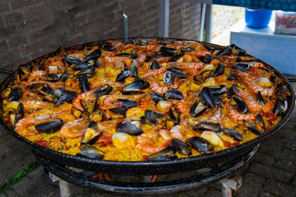 A massive, shallow pan filled with vibrant seafood paella sits outdoors on a metal stand. The dish is packed with saffron-infused rice and a generous assortment of large, whole shrimp and dark-shelled mussels.