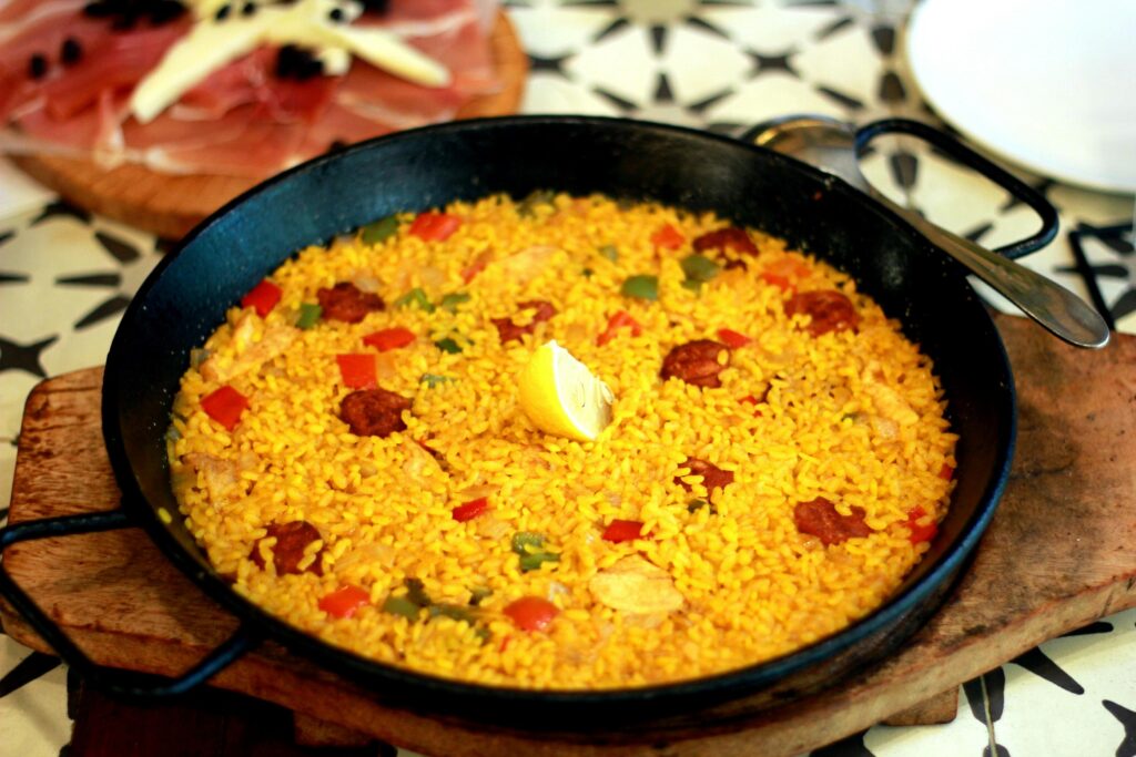 A vibrant paella featuring yellow rice, red peppers, and sliced chorizo is served in a black pan with a single lemon wedge at the center. The dish sits on a rustic wooden board, accompanied in the background by a plate of cured meats and cheese.