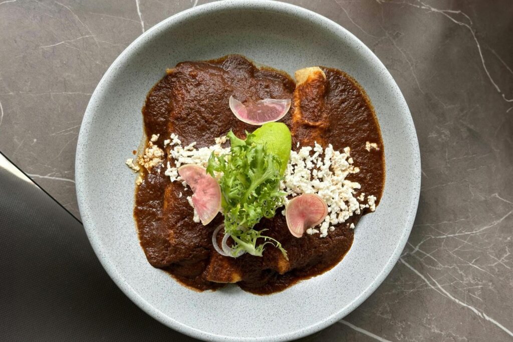 This dish features two enchiladas smothered in a rich, dark mole sauce and served in a light grey textured bowl. The plate is elegantly garnished with crumbled white cheese, fresh greens, and thinly sliced radishes.