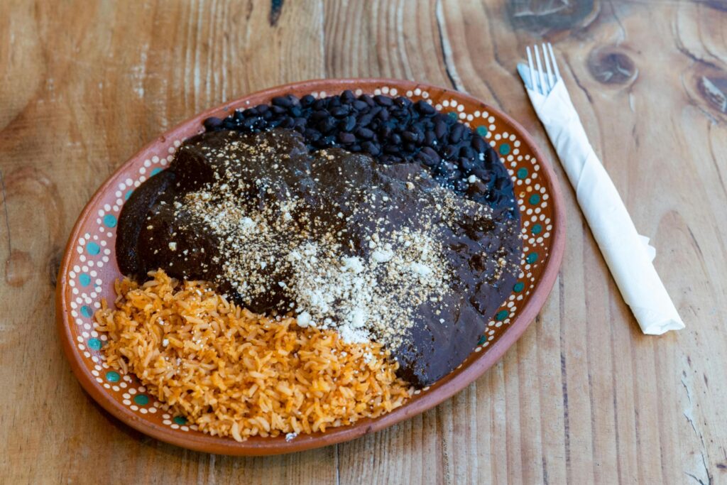 This traditional Mexican dish features a protein, likely chicken or turkey, completely covered in a thick, dark mole sauce and dusted with sesame seeds or crumbled cheese. It is served on a decorative clay plate alongside portions of orange-tinted rice and whole black beans.
