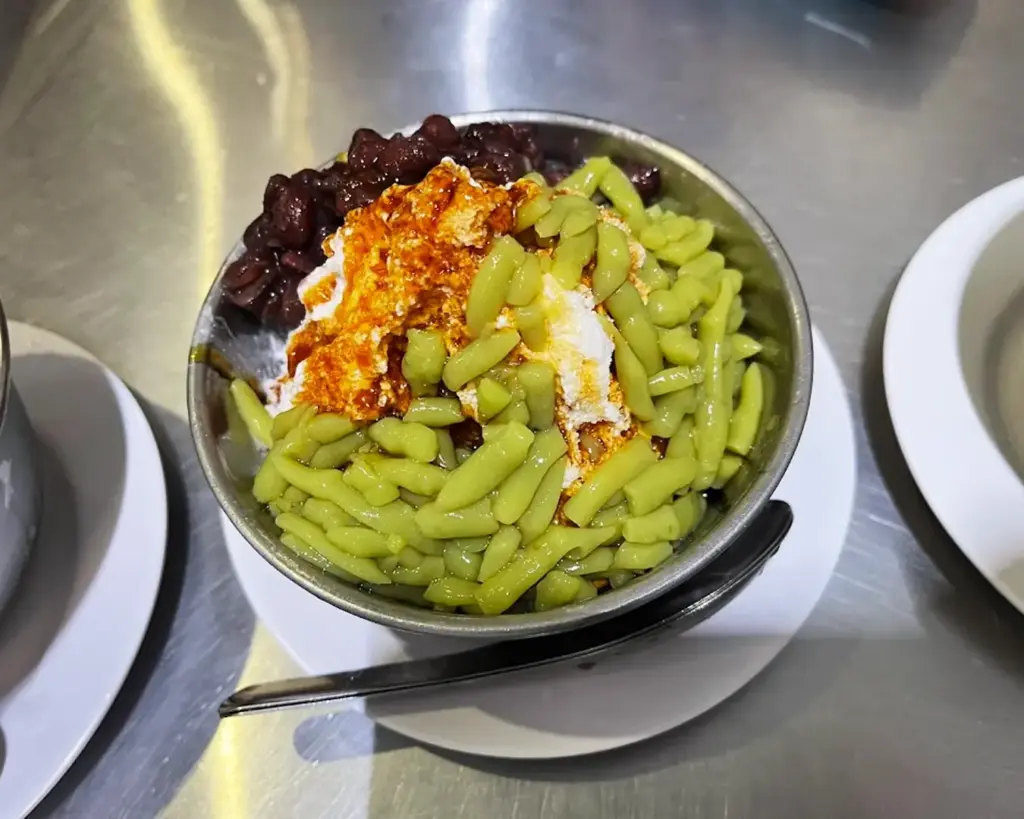 A bowl of cendol from Yat Ka Yan Dessert, filled with shaved ice, coconut milk, palm sugar syrup, and green jelly strands, creating a refreshing and creamy Southeast Asian dessert.