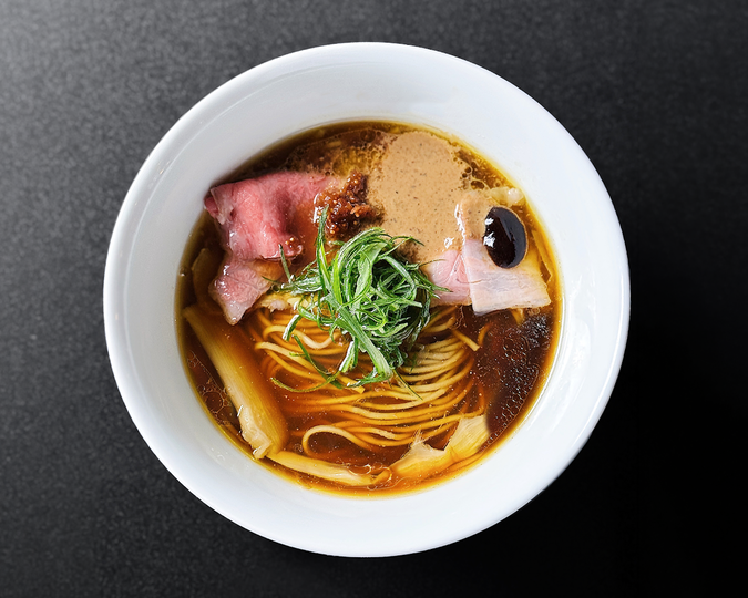 A bowl of ramen with thin noodles in brown broth, topped with slices of meat, green onion, and a dark sauce, is presented on a dark surface.