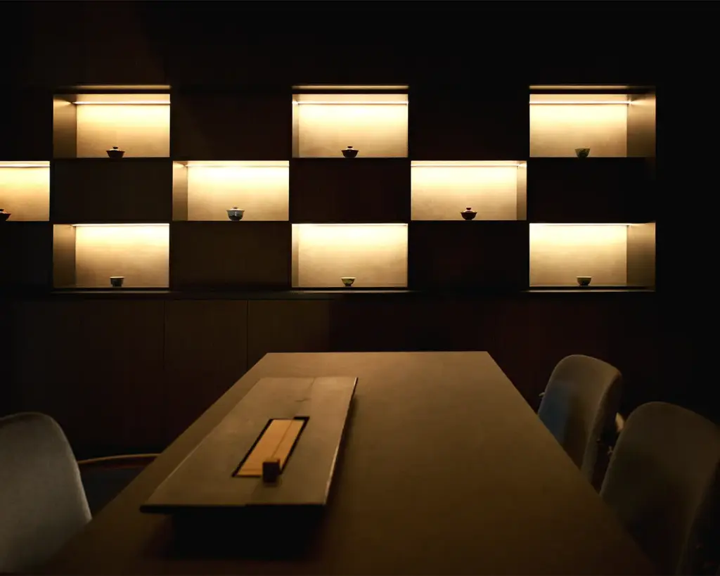 Dimly lit minimalist room featuring a wooden table and illuminated shelves displaying small bowls, creating a warm, serene ambiance.
