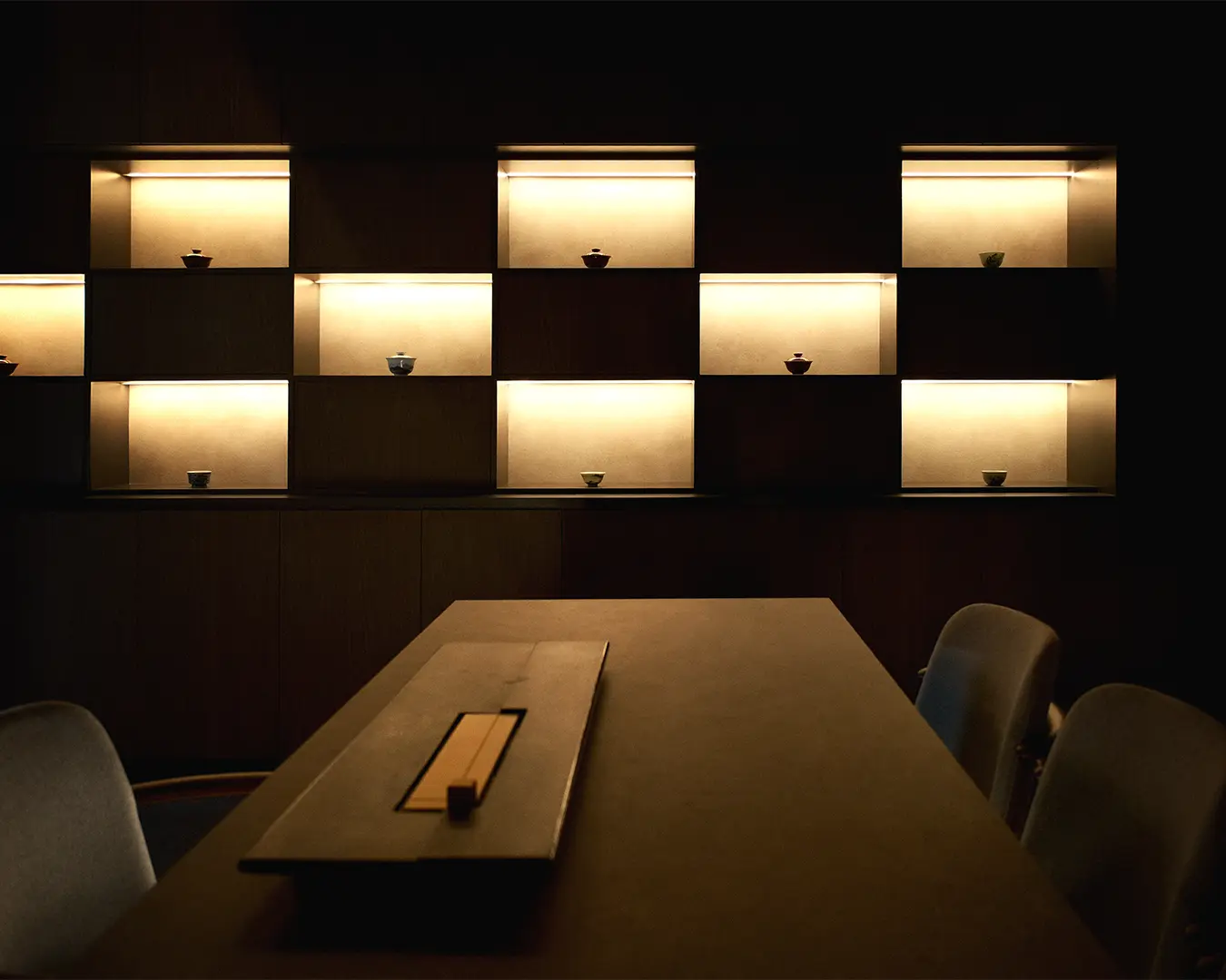 Dimly lit minimalist room featuring a wooden table and illuminated shelves displaying small bowls, creating a warm, serene ambiance.