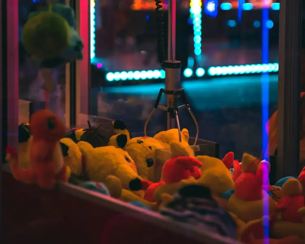 Arcade claw machine filled with colorful plush toys, including yellow and red figures. Neon lights create a vibrant, playful atmosphere.