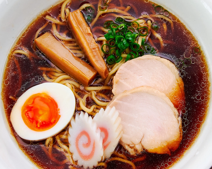 A bowl of ramen with dark broth, topped with sliced pork, green onions, narutomaki, bamboo shoots, and a soft-boiled egg with a runny yolk.
