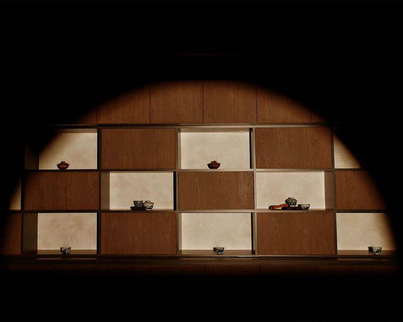 Dimly lit wooden shelf with a geometric pattern, displaying small bowls and cups. The spotlight creates a warm, serene ambiance.